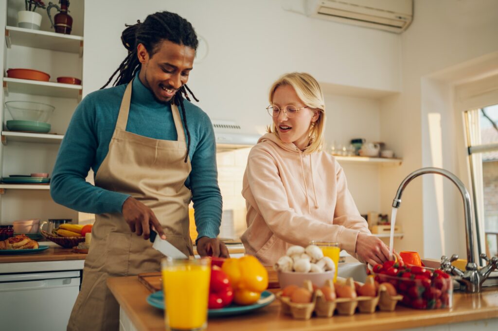 A cheerful interracial couple is preparing lunch and doing dishes at home.