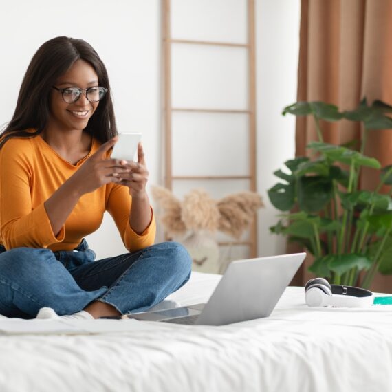 African American Woman Using Phone And Laptop Sitting In Bedroom