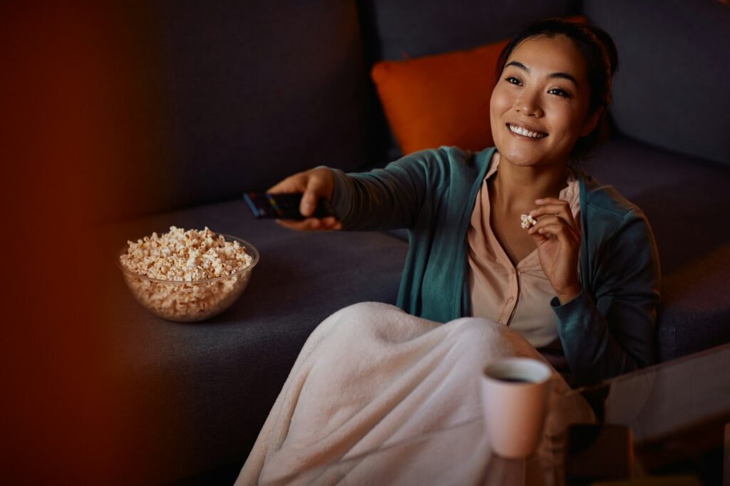 Happy Asian woman eating popcorn while watching TV at home at night.