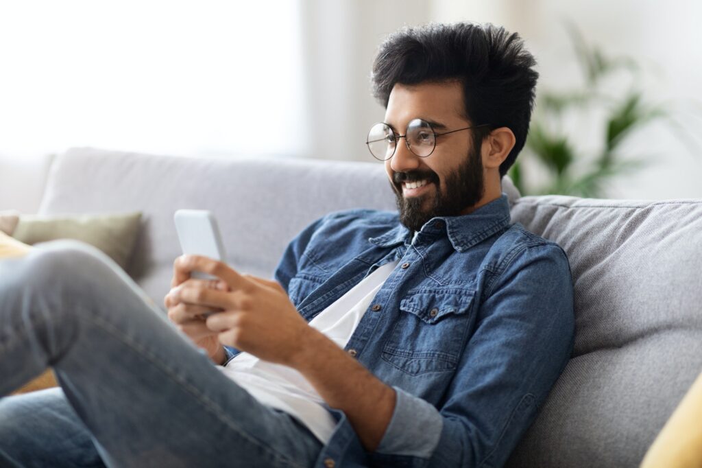 Dating App. Joyful young indian man relaxing with smartphone at home