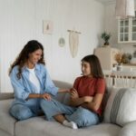 Shy introverted teenage girl sits in closed position on couch with mother wanting to play together