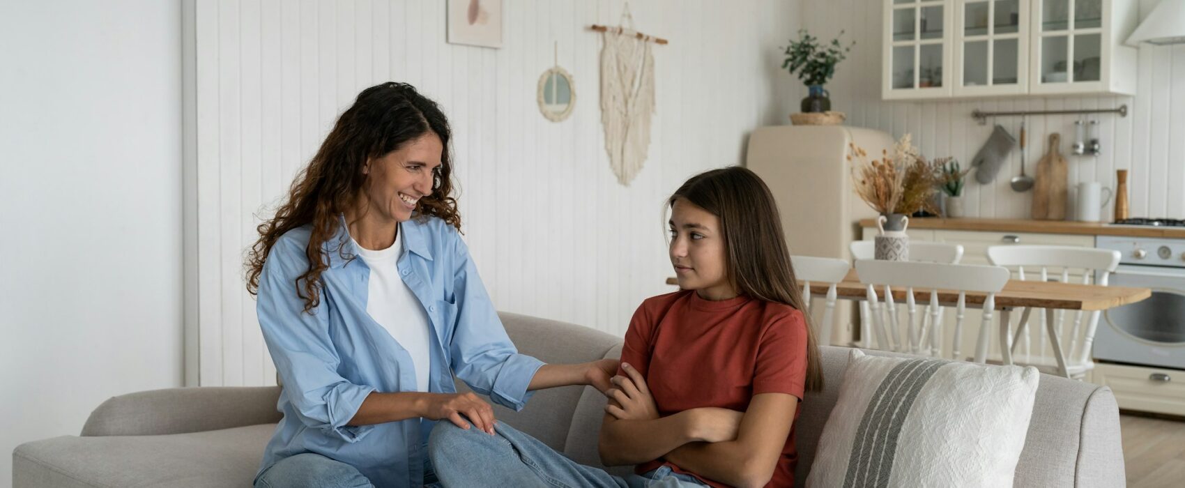 Shy introverted teenage girl sits in closed position on couch with mother wanting to play together