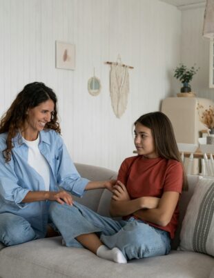 Shy introverted teenage girl sits in closed position on couch with mother wanting to play together