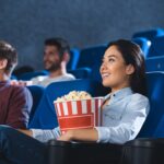 smiling asian woman with popcorn watching movie in cinema alone