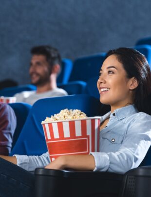 smiling asian woman with popcorn watching movie in cinema alone