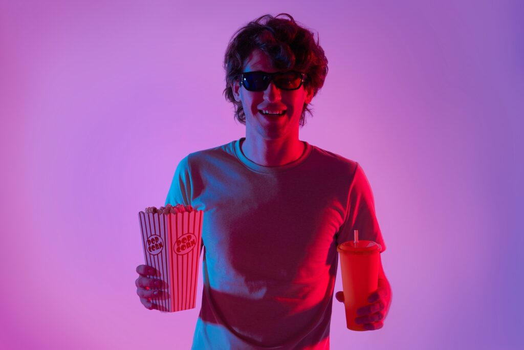 Young man wearing 3D glasses enjoying eating popcorn and watching movie on neon lights background.