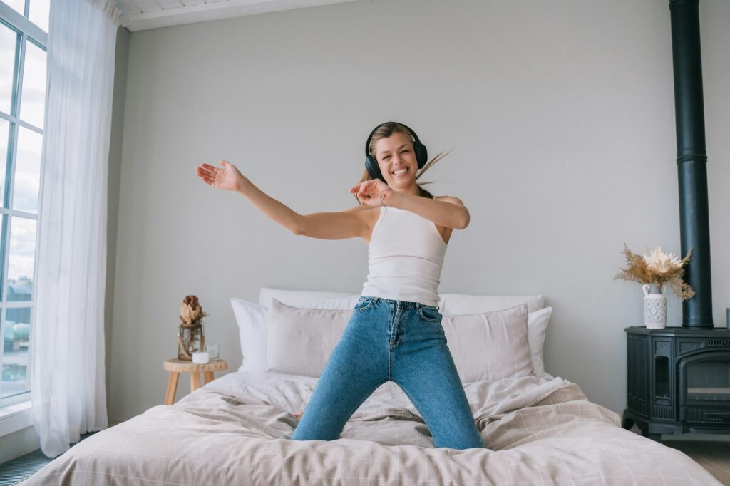 cheerful young woman dancing on bed, enjoying music with headphones, in a bright, airy bedroom