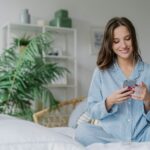 Happy woman with appealing look, holds modern cell phone, downloads song to playlist