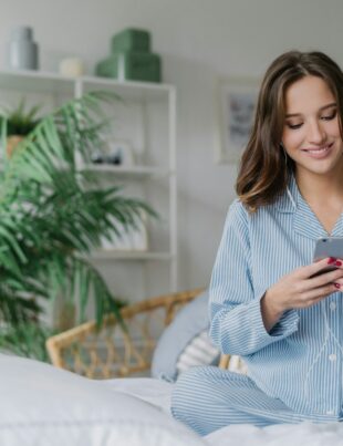 Happy woman with appealing look, holds modern cell phone, downloads song to playlist