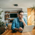 Portrait of a casual businessman thinking while looking at laptop screen, keeping hand on chin.