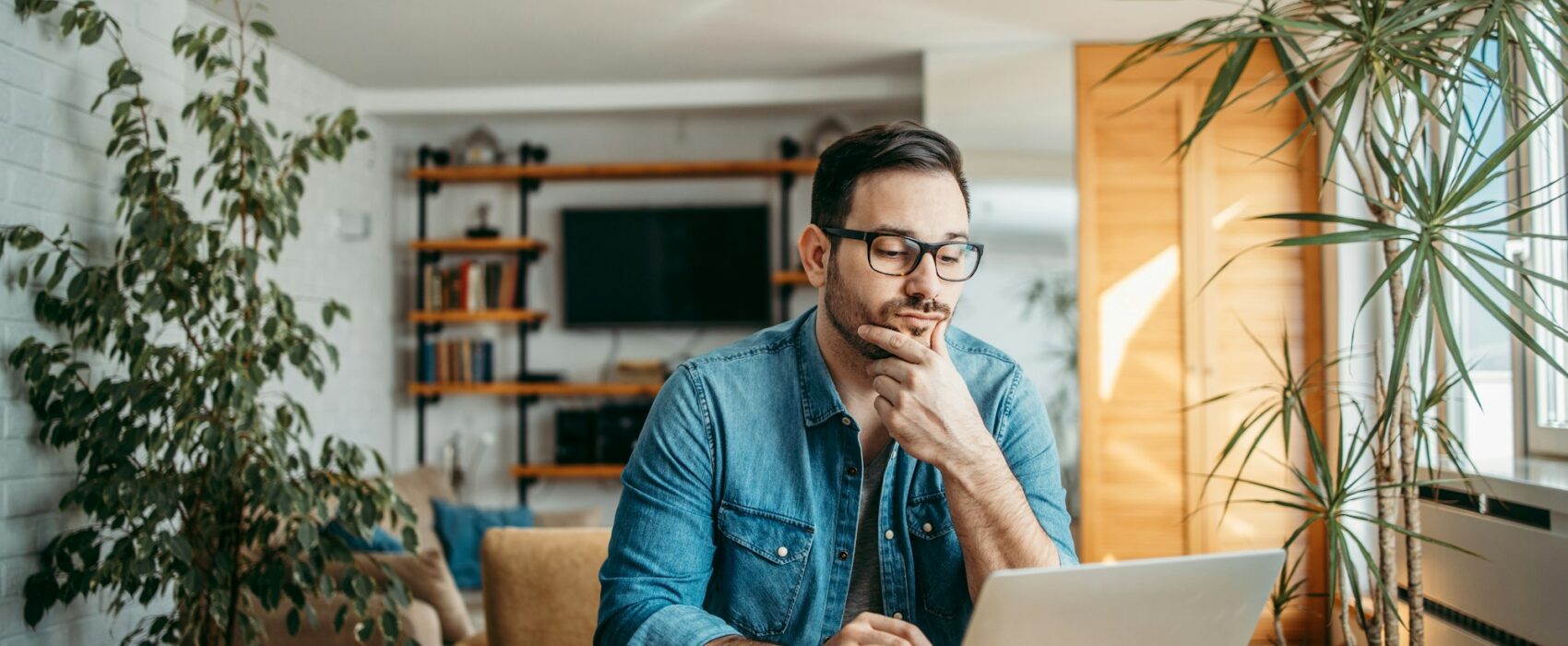 Portrait of a casual businessman thinking while looking at laptop screen, keeping hand on chin.