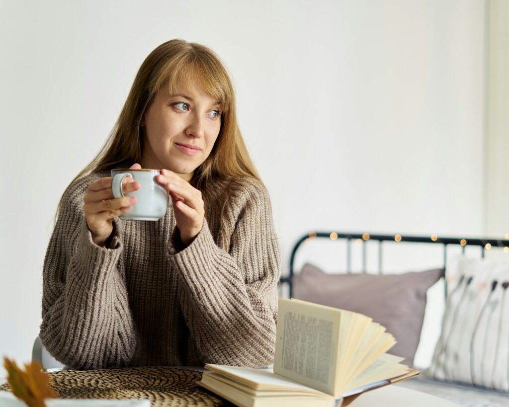 Happy young girl in sweater dreaming with cup of tea while reading book in bedroom